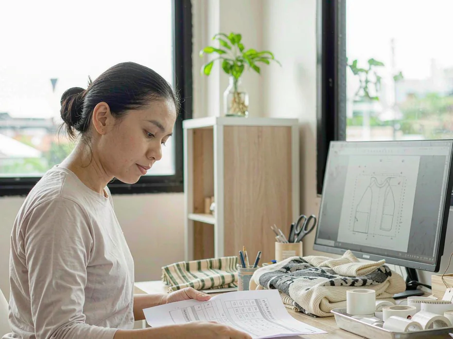 Merchandiser at a desk reviewing an order tracking sheet, with label artwork on screen and woven labels and hangtags in a tray.
